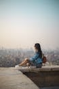A young woman in casual attire sits on a rooftop terrace, viewing a sprawling cityscape in daylight.